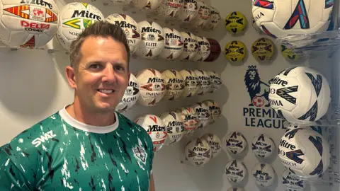 BBC Simon Francis is stood at the top of his stairs wearing a green with black and white stripes and smiling straight at the camera. Behind him are a large amount of Mitre footballs mounted and displayed above a staircase on a plain white wall. Each ball has a different Mitre model and design. Stuck at the centre of the back wall is a large Premier League logo. 