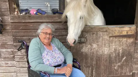 Jenny Morgan sits in a wheelchair wearing a duck egg cardigan and floral blue top with jeans. She has short grey hair and glasses and is sat next to a stable where you can see her grey horse peeping over the door.