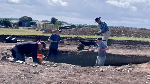 Four men around a big excavation dug out for an archaeological dig in the Borders. Two are using shovels to take dirt out of the hole, one has a wheelbarrow and the other is on his hands and knees next to an orange bucket. 