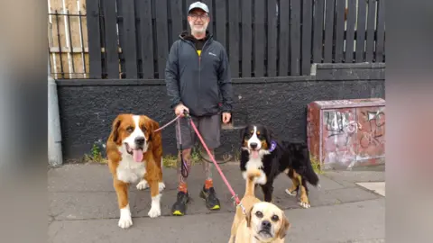 A man and his three dogs are looking are the camera. The man is wearing a cap and a navy jacket with grey shorts and trainers. Two of the dogs have their tongues out. 