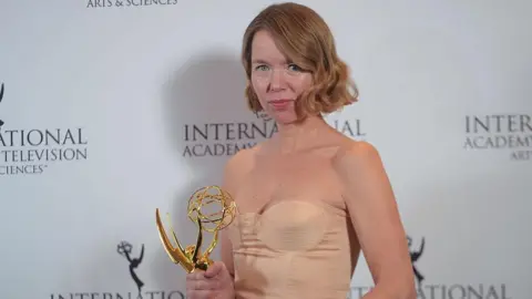 Getty Images Anna Maxwell-Martin stands in front of a backdrop covered in International Emmy branding. She is holding a large, shining gold trophy and is wearing a pale peach-coloured dress. She is looking directly at the camera, with a slight smile on her face.