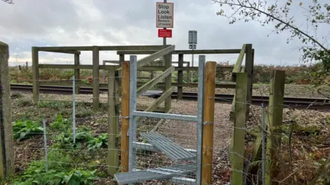 Vikki Irwin/BBC The picture is of railway crossing at Levington Heath which can only be accessed by foot. In the foreground you can see a metal stile with two steps and then the crossing is fenced off. There is a stop Look and Listen sign and the area is surrounded by countryside. 