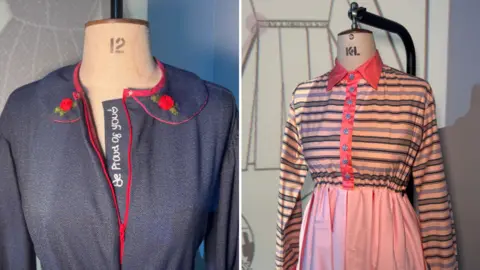 BBC/ Emily Johnson Two dress forms side by side: one wearing a dark blue dress with red piping, small embroidered red flowers on the collar tips and the words “Be Proud of You” along the neckline; the other wearing a dress with a pink skirt and a striped top in pink, blue and beige, finished with a bright pink collar and flower‑shaped buttons.