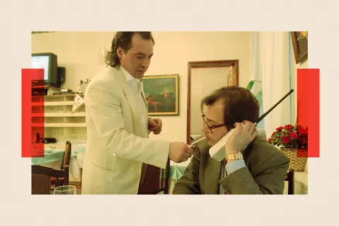 Getty Images A waiter in a white jacket lights the cigarette of a man sitting at a table holding a large old-style mobile phone
