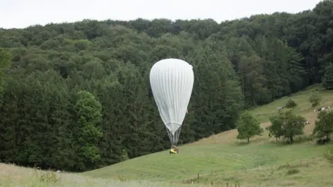 Johnny Green/PA A tall white hot air balloon lands in a hilly area of grass and forest