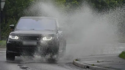 PA Media A black Range Rover drives through a large puddle on the side of a road during a rainy day, sending water flying high into the sky.