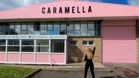Caramella Mollie-May Rossi is standing in front of a building which has been painted pink. A sign at the top of the building reads: Caramella. She has her back to the camera and is holding her right arm in the air. 