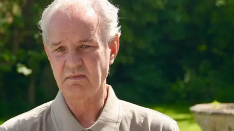 Ant Saddington/BBC A man with short grey hair, wearing a beige top with a collar. He looks at the camera as he sits outside on a sunny day. He is slightly frowning. Behind him is a garden, enclosed by trees.