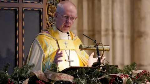 Archbishop Justin Welby speaks at the Canterbury Cathedral lectern, wearing a golden clerical robe speaking at a decorated pulpit adorned with Christmas greenery and ribbons inside a cathedral.