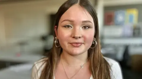 Martin Giles/BBC Beatrice looks straight at the camera. She has long, brown hair. She has hoop earrings and a ring through her nose. She is wearing a light coloured top. She is wearing a necklace.