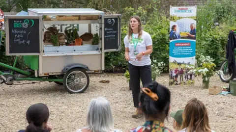Eden Project Communities The back of people's heads as they watch a talk given by a member of staff at the Eden Project. They are standing outside and there is a stall behind them on wheels, serving soup
