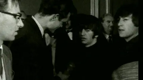 Ringo Star and John Lennon meet the press at the venue, then known as the ABC, when they performed there. This black-and-white image comes from BBC video footage. It shows Ringo in the centre of the image with distinctive sideburns. Jon Lennon is to his right.