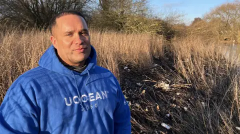 BBC Chris stands infront of rubbish strewn alongside the river at watermead country park 