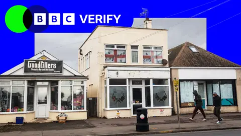 A photo of three small commercial buildings in Jaywick, near Clacton-on-Sea in Essex. The left unit is a sandwich shop named “Goodfillers,” the central building displays England flags in the windows above a tattoo studio, and the shop on the right has papered-over windows and a faded sign. Two men in are walking past, wearing dark clothing. The sky is grey and overcast. A BBC Verify logo is superimposed on the upper portion of the image with a blue border.