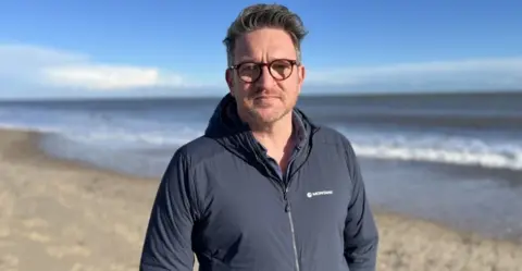 Shaun Whitmore/BBC Man with glasses looking directly at the camera while standing on a beach. he is wearing a blue jacket.