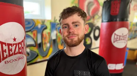 BBC Reuben Muston, a man with slightly curly brown hair and a beard, is pictured in the gym. He is stood in front of two large punch bags, behind which is a wall with graffiti on it. 