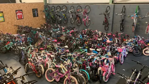 Hundreds of bikes are lined up in a warehouse of all different colours and sizes. A number are also hung on the wall at the back of the room. 