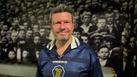 El aficionado al fútbol Kevyn Whitelaw, con una camiseta de Escocia, sonríe con el telón de fondo de un enorme mural de aficionados escoceses en Hampden Park.