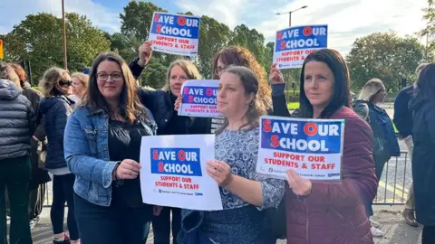 PA Media Five women outside school gates holding pieces of paper say 'Save Our School'