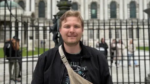 Henrick is standing outside railings, behind him a grand looking white washed Belfast City Hall. He has blond curtains in a middle parting and blond stubble. He is wearing a black t-shirt and jacket with a cream banana bag on top. He is smiling at the camera. 