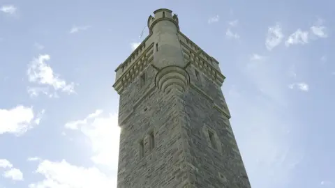 A view up towards the top of the MacDonald Monument. It is a tall, stone tower with narrow windows, and faux turrent and battlements. It is a sunny day.