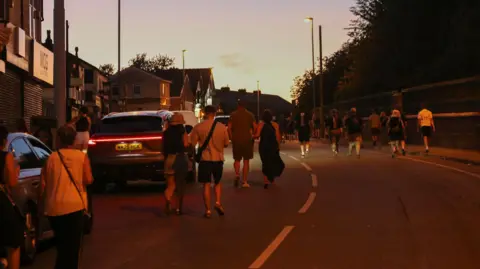 Emma Gibbs People in shorts and T-shirts walk up the road in the late evening light after Oasis at Heaton Park