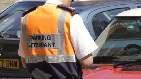 A parking attendant wearing a high visibility jackets and white shirt stands in front of two parked cars with his back to the camera