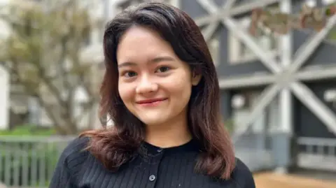 ऑड्रे हिल-विकावा, 20, काळा कार्डिगन घालते आणि तिच्या खांद्यापर्यंत काळे केस आहेत. तो एका झाडासमोर आणि इमारतीसमोर उभा राहतो आणि कॅमेरासाठी हसतो