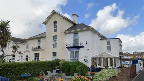 The Holly-Lets building is a mix of two and three storeys with a curved sign reading HOLLY LETS. One of the windows at the top is arched and another has an iron balcony. To its left is a two storey  building with a blue  sign reading SEASIDE GUESTHOUSE. One of its windows also has an iron balcony. There is a round conservatory on the side and a garden with tables and chairs and a palm tree, surrounded by a high hedge. A car park and the road are just visible on the left.