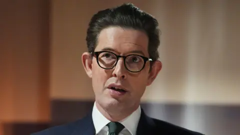 Getty Images Sir Ken McCallum, a white man with dark hair swept back and dark-rimmed glasses. He is wearing a dark suit with a white shirt and a dark tie and is photographed with a telephoto lens as he appears to be speaking at an event