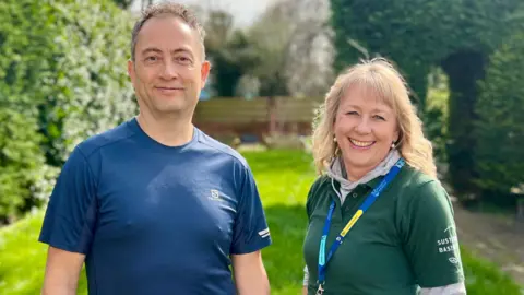 A man and woman are in the centre of the image. The man on the left is wearing a blue t-shirt. The woman on the right is wearing a green t-shirt and a lanyard around her neck. She is an eco expert from Basingstoke and Deane Borough Council and is visiting the man's garden to offer advice on how to make it more wildlife friendly.