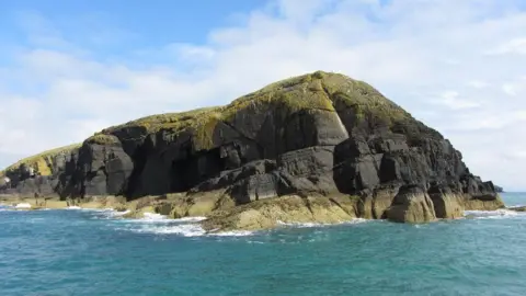 Geograph/Gareth James A view looking at the island from the sea. The rocky island rises up to a high cliff with rocks also jutting out into the sea, and greenery on top. There is a blue sky with white clouds, and the sea is turquoise. 