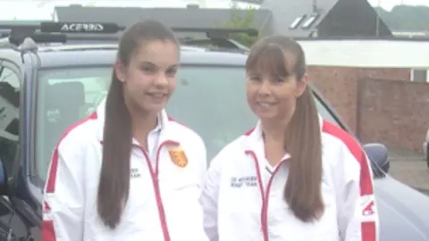 Amy Dingle Amy and her mum Karen are standing side by side in matching sports tracksuits, wearing white jackets with red accents and red trousers. 