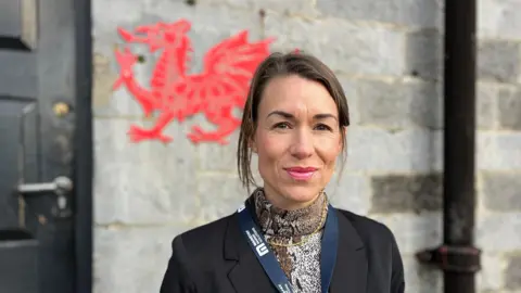 A woman with tied-back brown hair wearing a animal print blouse and black blazer. She is looking at the camera and smiling with a wall behind her, on which is a red Welsh dragon. 