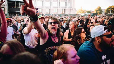 Nadine Ballantyne A large group of fans are packed into the Lloyds Amphitheatre during Bristol Sounds to watch the band Skindred. Many of them are looking towards the stage but one man is facing the camera and holding out an arm as he sings along. He has a black vest on, a goatee and moustache and sunglasses.