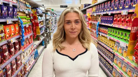 Beth standing in a supermarket aisle lined on both sides with Easter eggs. She has blonde wavy hair, shoulder length, and is wearing a silver necklace and a cream v-necked ribbed top with black edging around the neck.