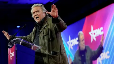 Steve Bannon in the midst of a speech in front of a large illuminated screen, waving his arms