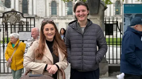 A man and a woman are standing outside railings with a brown pram, behind them is a grand looking white washed Belfast City Hall. An elderly couple walk behind them - the elderly lady has a yellow coat on.
The woman is wearing a beige cream trench coat over a black top and she has brown long hair with sunglasses on her head and earrings. She is smiling and has her hands clapped resting on her brown pram.
The man has a dark grey puffer jacket on and has his hands in his pockets. He has brown longish curtains and he is smiling at the camera.