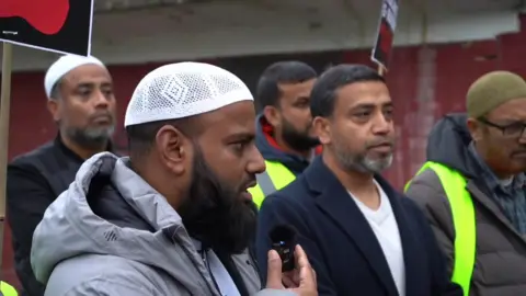 Darul Arqam Masjid and Muslim Community Centre Men taking part in a "walk of solidarity", they are holding up signs and a bearded man closest in the picture is wearing a white Muslim prayer cap and is speaking into a small microphone