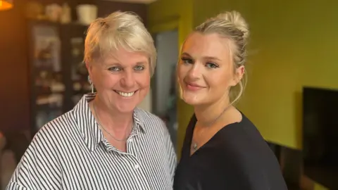 Women with blonde hair wearing a black and white striped shirt alongside a younger woman with blonde hair wearing a black longsleeve top.