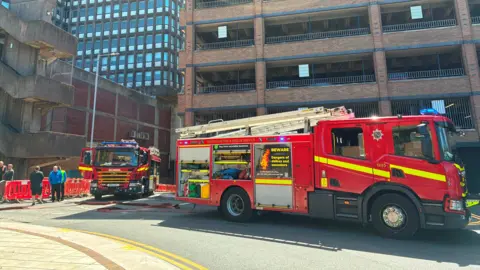 Two fire engines outside a car park near to the Strand shopping centre in Bootle. Several people are stood next to red barriers.