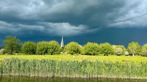 Weather Watchers / Katrina River in front, with reeds on the side, a green ,meadow and trees behind with a flock of grazing sheep. Sun bounces off the green with the cathedral spire in the distance. However, the clouds overhead are a deep grey.