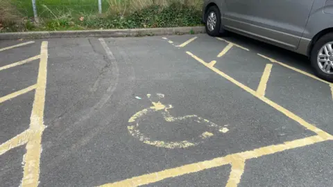 LDRS A disabled parking bay is marked out in yellow paint. A silver car is parked in the space next to it. There is a kerb marking the car park boundary with a fence and grass in the background.