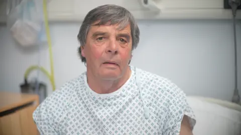 Jamie Niblock/BBC Trevor Wilson with short hair wearing a hospital gown sitting up in a hospital bed.