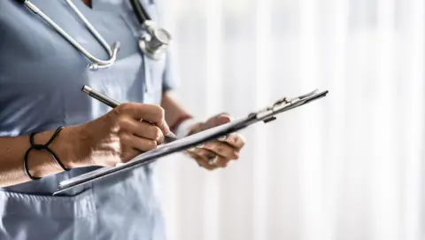 A woman in light blue nurses scrubs has a stethoscope around her neck. She is writing on paper on a clipboard.