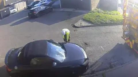 Black car sits in a large bay marked with white lines drawn around it. The car sits in a cul-de-sack with other vehicles. A contractor works on the ground. 
