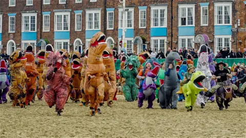 We Are Weymouth A group of people wearing dinosaur costumes running along a beach. Spectators are watching from a promenade behind them, with a row of brick buildings behind them.