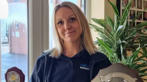 Pave-Aways A woman with blonde hair holds up trophies in an office reception area. She is smiling and wearing a navy polo-shirt with PaveAway company logo just seen, and fleece. A plant can be seen behind her and a window on to a carpark.