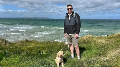 Scott Adams Scott Adams wearing grey raincoat and shorts, standing on grass in front of ocean with his dog. 