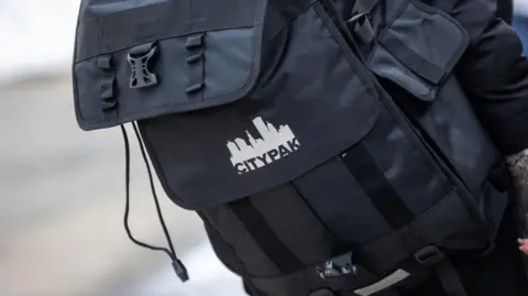 PA/CITYPAK Large black rucksack with CITYPAK logo on it. The rucksack has plenty of large compartments and the background is blurred. 